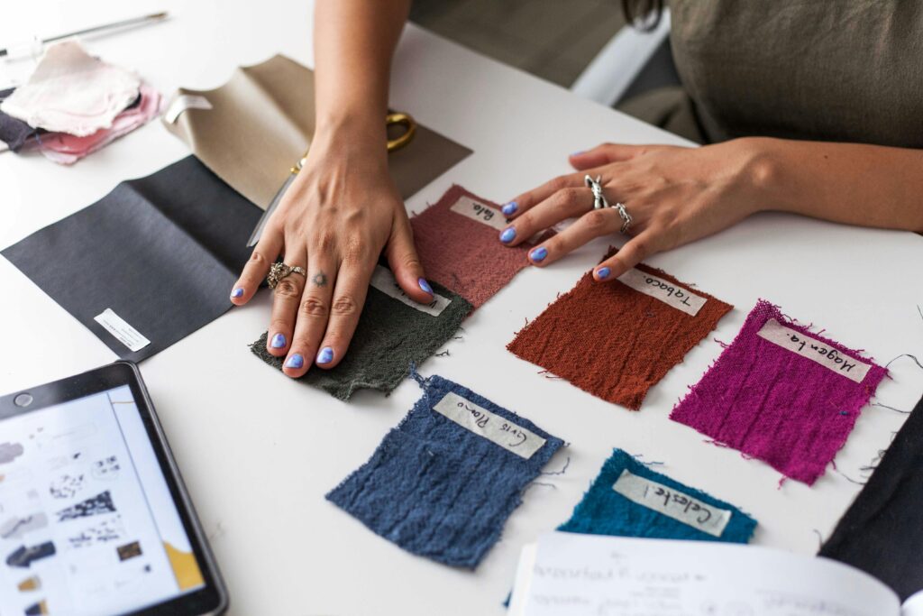 A person with painted nails examines colorful fabric swatches labeled "Blue Mel" on a white table, with a tablet and notes nearby.